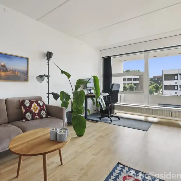 En stue med en brun sofa ved siden af en gulvlampe, et lille rundt træbord foran, plante ved siden af. Skrivebord og stol ved et stort vindue viser lejlighedsbygninger udenfor.