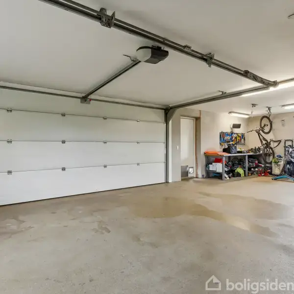 En rummelig garage med en stor hvid port. Cykler hænger på væggen og står ved siden af hylder med værktøj. Gulvet er beton, og der er et vindue mod højre.