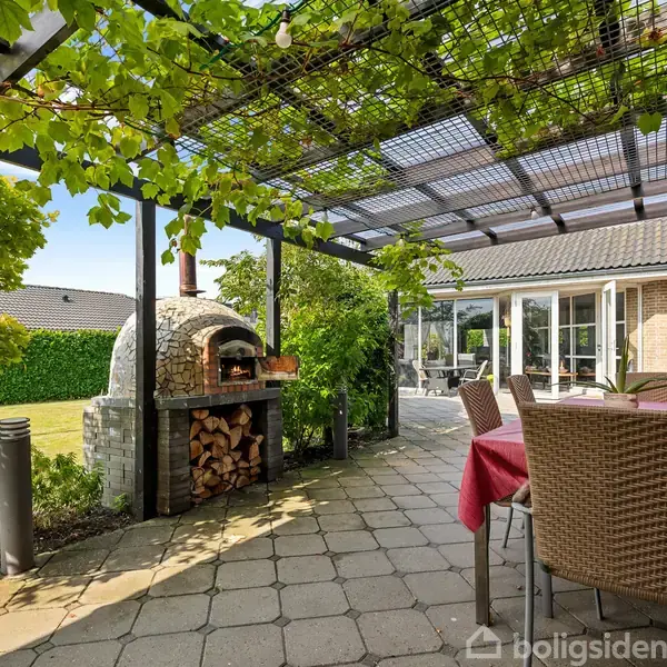Udekøkken med kuppelovn, placeret ved siden af et bord med rødt dug under en pergola dækket af vinstokke. Græsplæne og buske omkranser terrassen i et solrigt haveområde.
