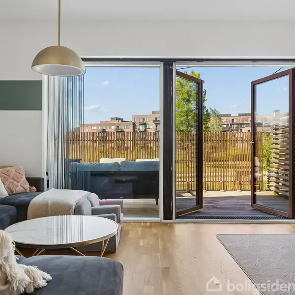 Sofa med puder og tæppe står i stue ved siden af et rundt bord. Dobbeltglasdøre åbner til en balkon med udsigt til bygninger og træer.