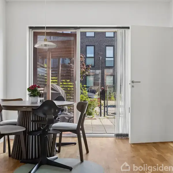 Et rundt spisebord med fire stole står i et lyst køkkenområde. En dør og store vinduer fører ud til en terrasse med udsigt til en moderne bygning.