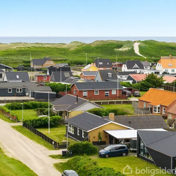 Huse med farverige tage står tæt i en kystnær landsby. En grusvej og græsarealer omkranser boligområdet. I baggrunden ses en klitrække med en sti, der fører mod havet.
