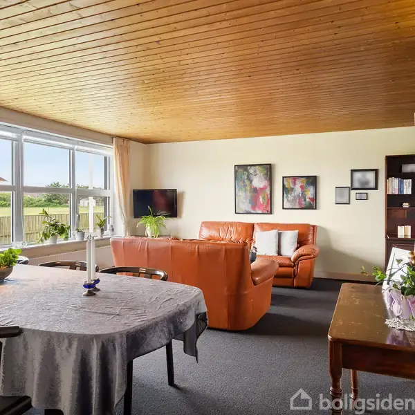 Stue med brun lædersofa og sofabord, indrammet af store vinduer og lyse vægge. Spisebord med dug og planter er i forgrunden. Træbeklædt loft og dekorationer på væggen.