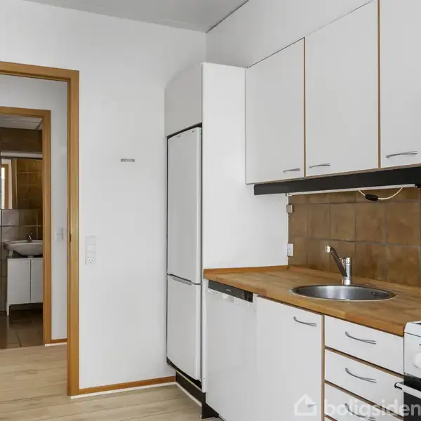 Køkken med hvide skabe og træbordplade; en vask monteret i bordpladen. I baggrunden ses en åbent dør til et badeværelse med toilet og vask. Ingen tekst fundet.