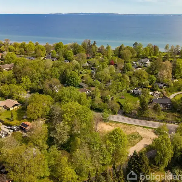 Kystnært boligområde med spredte huse omgivet af frodig grøn vegetation. Baggrunden domineres af en blå havhorisont, mens veje snor sig gennem området.