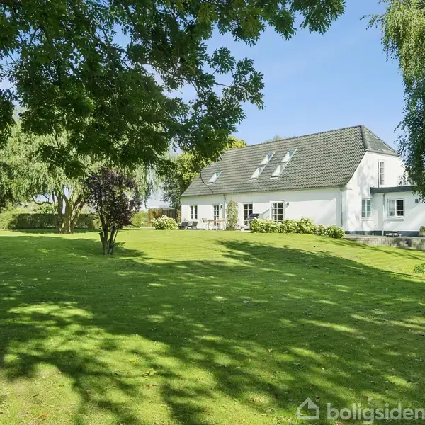 Hus med hvid facade og sort tag står i solrige omgivelser. En stor, grøn have med træer omgiver huset. Det er en fredelig og landlig scene.
