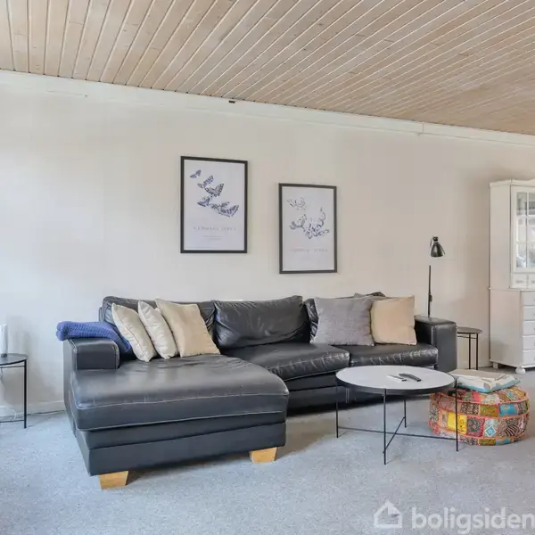 Sort lædersofa står i en stue, omgivet af sofabord og farverig puf. Væggen bagved har to billeder. Til højre ses en hvid skænk med glasvitrine.