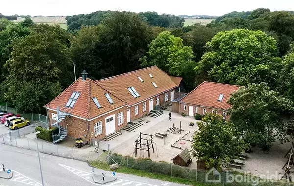 En murstensbygning med rødt tag står omgivet af træer. To legepladser med klatrestativer ligger foran bygningen. Bilen står parkeret ved vejen til venstre.