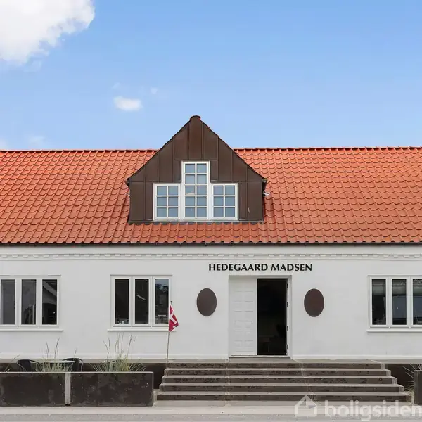En hvid bygning med rødt tegltag, tekst "HEDEGAARD MADSEN" over hovedindgangen placeret i midten, omgivet af trapper og en vej. Nummer 55 ses på højre side.