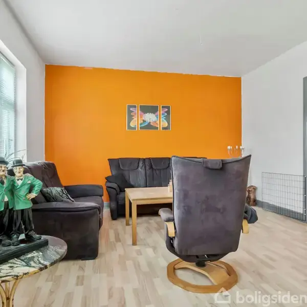 En stue med en sort lædersofa og en lænestol arrangeret omkring et trækaffebord. En lys orange væg dominerer baggrunden. På sidebordet står to dekorative figurer.