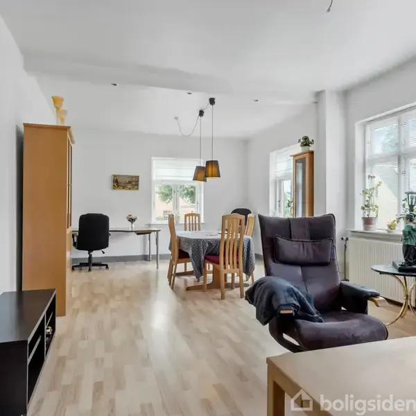 Stue med trægulv og hvide vægge, møbleret med sofagruppe og spisebord. Vinduer med planter giver naturligt lys. Sorte og træfarvede møbler dominerer rummet, der er lyst og indbydende.