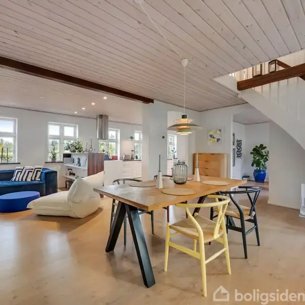 Spisebord med stole står i midten af et lyst, moderne stueområde. Til venstre er der en sofa ved vinduerne, og en trappe fører opad til højre.