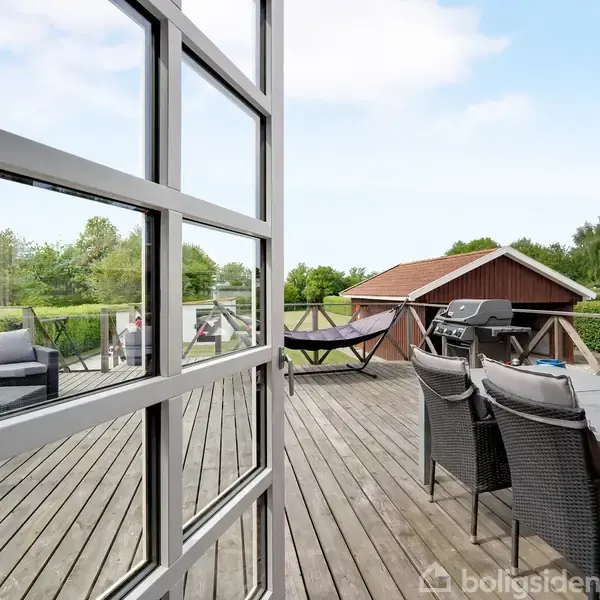 En terrasse med havemøbler og en grill står på et trædæk. En hængekøje hænger ved siden af et rødt skur. Grønne træer og buske omkranser området.