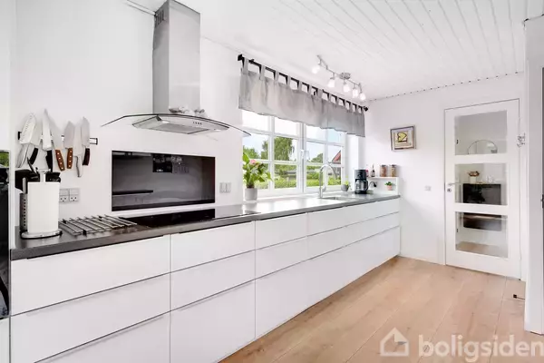 Moderne køkken med hvidt skabsarrangement, glaskomfur og emhætte; store vinduer lader dagslys strømme ind. Lys træparket og pæne dekorationer skaber en harmonisk atmosfære.