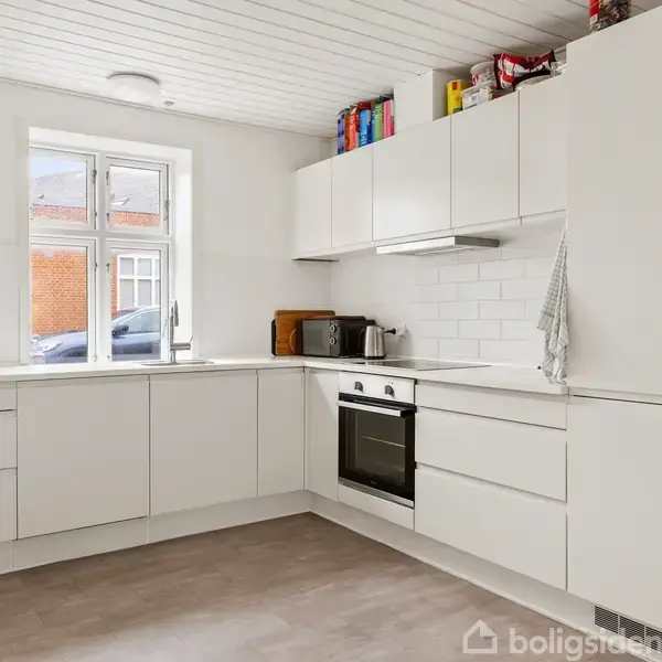Et hvidt køkken med moderne skabe arrangeret i en vinkel. En ovn er integreret i skabenheden, mens en kaffemaskine står på bordet. Et vindue giver naturligt lys.