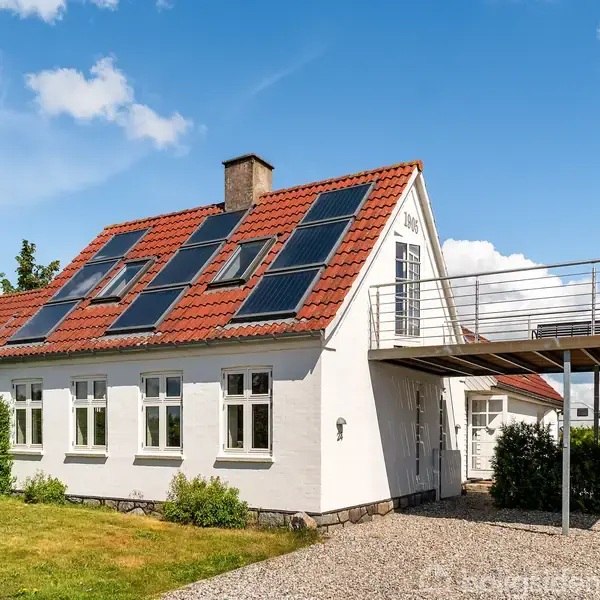 Et hvidt hus med rødt tag har solpaneler monteret. En grussti leder til indgangen. Omkring huset er der en velplejet have og en balkon. Skyer og blå himmel ses i baggrunden.