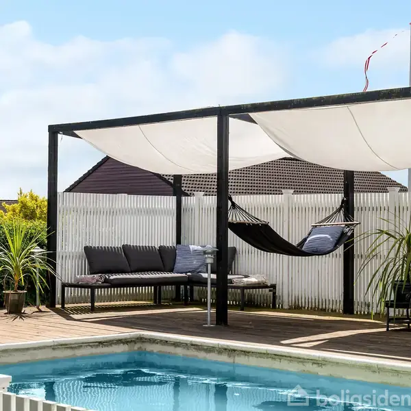 En sort hængekøje hænger under en pergola med hvidt solsejl. Ved siden af er en sofa på en terrasse ved en swimmingpool. Der er planter og en hvid plankeværk omkring.