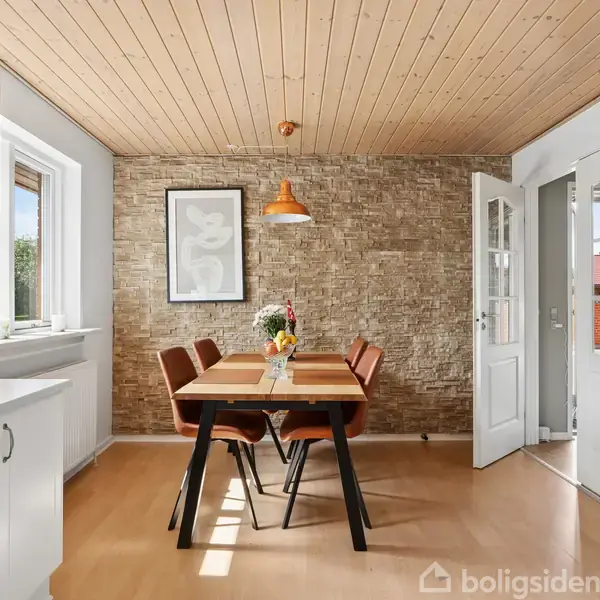Et spisebord står centralt i en lys spisestue med blomster på bordet og fire brune stole omkring. Omgivet af træloft, murstensvægge og store vinduer.