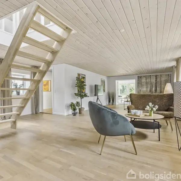 Trætrappe fører op til en øvre etage i en rummelig stue med hvide vægge og trægulve. Rummet indeholder moderne møbler og vinduer med lysindfald gennem store gardiner.