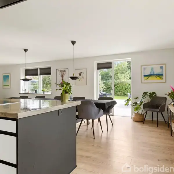 Et moderne køkken-alrum med et stort køkkenbord i forkant. Spiseplads med sorte stole og grønne planter. Rummet har hvide vægge og store vinduer med udsigt til haven.