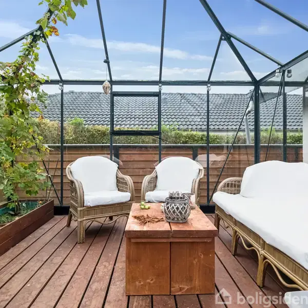 Et drivhus med glasvægge, der indeholder kurvemøbler med hvide hynder. Et træbord står i midten. Til venstre vokser tomatplanter i en plantekasse.