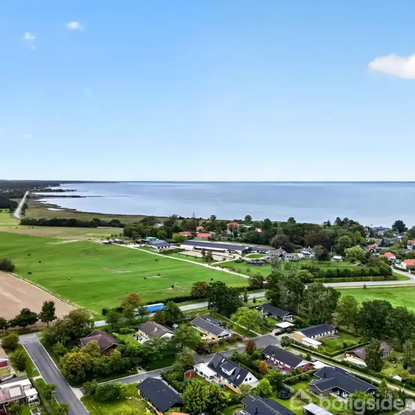 Kystudsigt strækker sig over grønne marker og et boligområde med spredte huse. Havet breder sig langs horisonten under en klar, blå himmel.
