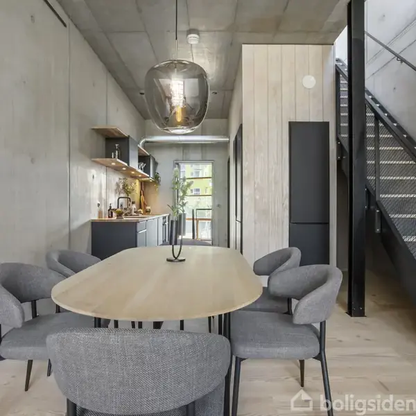 Et moderne køkken-alrum med en lang spisebord, omgivet af seks grå stole. Væggene er beton, og en metaltrappe fører til en anden etage. En stor lampe hænger over bordet.