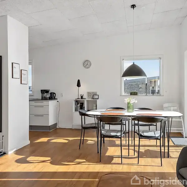 Et rundt spisebord med fire stole står i et lyst køkken-alrum. Sollys strømmer ind gennem en stor skydedør til højre. Der er en sort lampe over bordet.