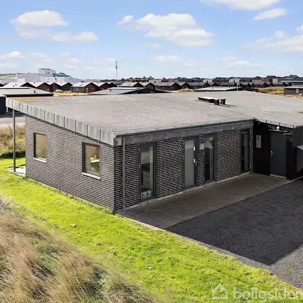 En moderne, sort murstensbygning står på en grusbelagt grund omkranset af grønt græs. Bygningen har store vinduer og er beliggende i et villakvarter under en lyseblå himmel med spredte skyer.