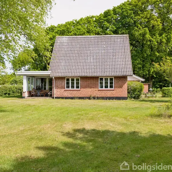Et rødt murstenshus med sort tag står i en grønt område omkranset af træer. Ved siden af huset er der en overdækket terrasse med møbler.