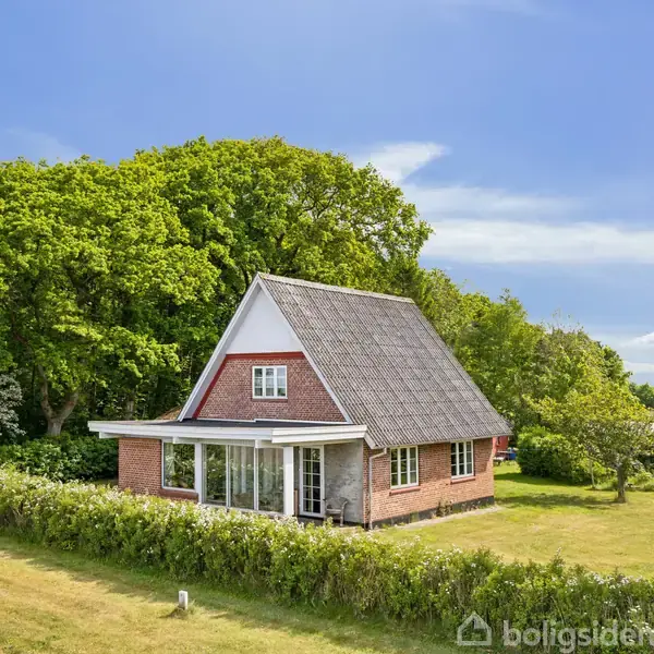 Et rødt murstenshus med et skråt tag står omgivet af en grøn have og tæt skov. En glaseret veranda ligger foran huset, og himlen er blå og klar.