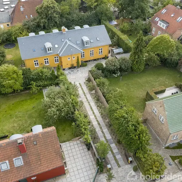 Orange hus med blå tag står omgivet af frodige haver og grønne træer, flisebelagt sti fører op til hovedindgangen. Nabohuse med røde og grønne tage omgiver.