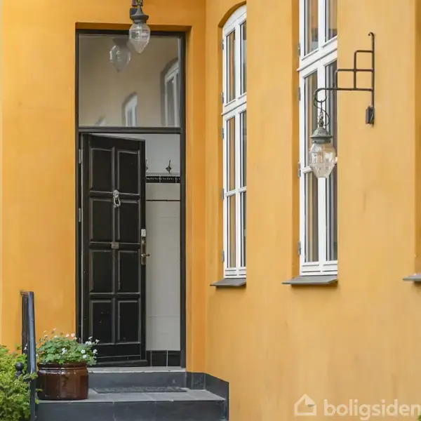 Sort trædør står åben ind til lys entré, flankeret af orange facade og hvide vinduer. En glaslampe hænger over indgangstrappen ved siden af grønne buske.