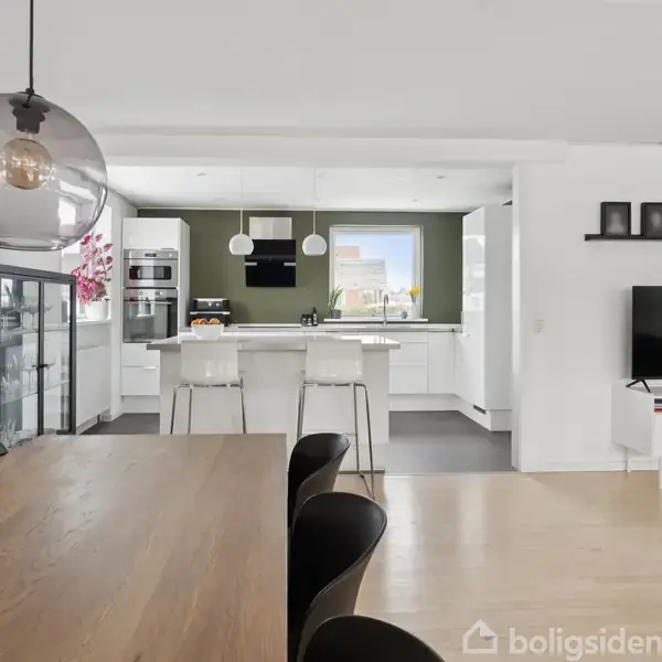 Lys og moderne stue-køkken i åbent plan. Et træbord med sorte stole står foran, mens et moderne køkken med barstole er bagved. En fladskærms-tv er placeret på en hvid skænk.