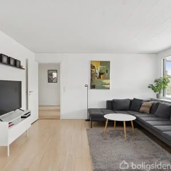 Stue med grå hjørnesofa og lille rundt sofabord på et gråt tæppe. Fladskærms-TV på hvidt møbel, hylde med dekorationer ovenfor, og store vinduer til højre.