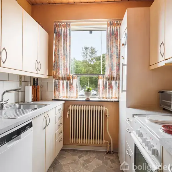 Et køkken med hvide skabe og fliser. Vasken og opvaskemaskinen er til venstre, komfur og mikrobølgeovn til højre. Et vindue med blomstrede gardiner viser et grønt landskab udenfor.