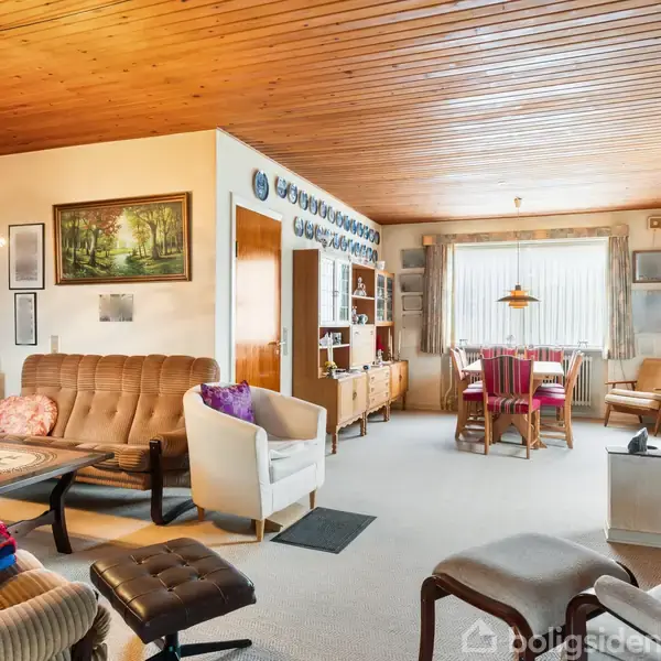 Stue med sofa, lænestole og sofabord står i forgrunden. I baggrunden ses et spisebord med stole under et stort vindue. Væggene er pyntet med kunst og keramik.