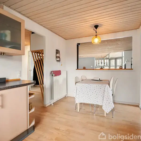 Spisebord med dug står i et lyst køkkenalrum, omgivet af hvide vægge og lyse trægulve. Et vindue og pendellampe skaber en hyggelig atmosfære.