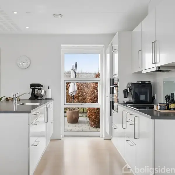 Moderne køkken med hvide skabe, to køkkenborde med apparater og redskaber. Lys strømmer ind gennem et stort vindue, og en terrassedør fører ud til en have med planter.