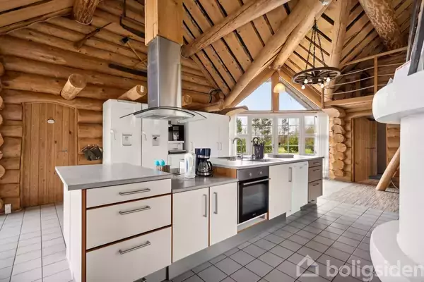 Køkkenø med hvide skuffer og indbygget ovn står midt i et rum med tømmerloft. Store vinduer fylder rummet med naturligt lys. I baggrunden ses en dør og en trappe.