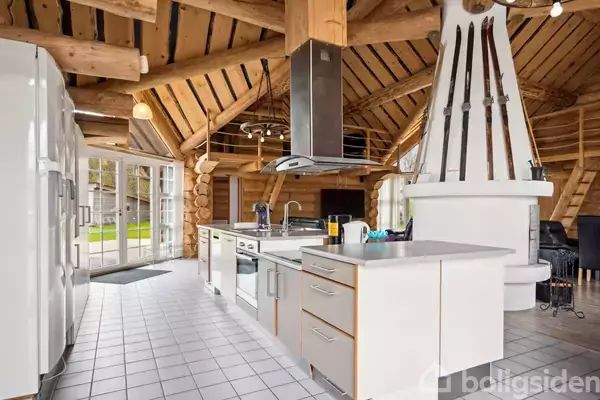 Et moderne køkken med hvidt modulisystem og en emhætte i rustfrit stål. Det har træbjælkelofter og store glasdøre, der åbner ud til en have. En pejs med dekorationer er i baggrunden.