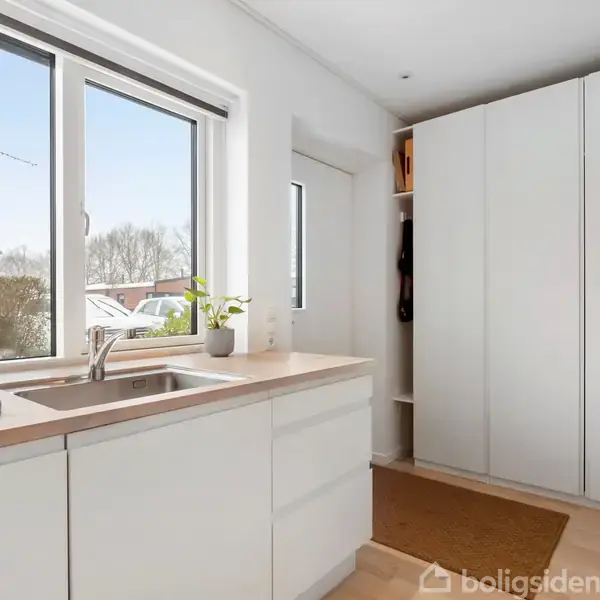 Køkken med hvidt skab og vask, brød og plante på bordpladen. Store vinduer viser et træ og et hus udenfor. Skydedør ved siden af skabet.