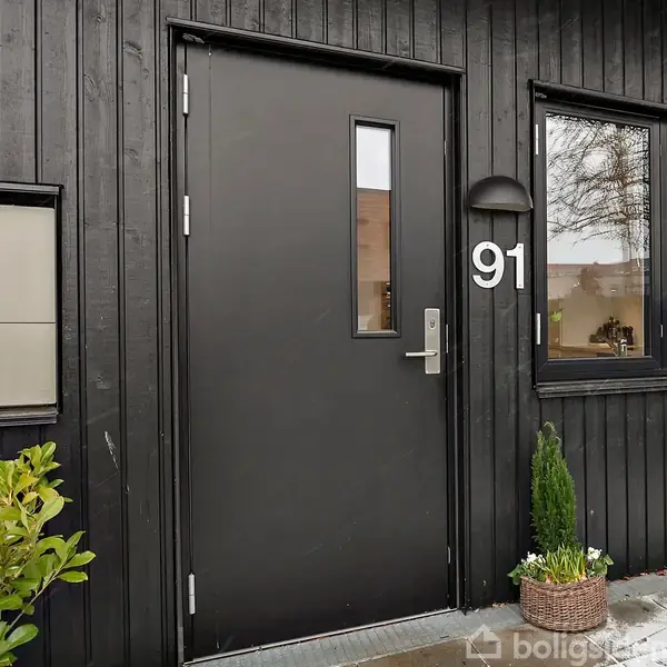 Sort hoveddør med et smalt vindue står i stilfuld, moderne træfacade. Til højre ses husnummeret "91" og et vindue. Foran døren står en lille plante i potte.