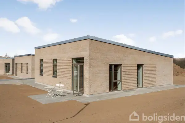 En moderne etplans murstensbygning med store vinduer står omgivet af en åben jordgrund. Foran huset er et lille udendørs bord med to stole placeret på fliser.