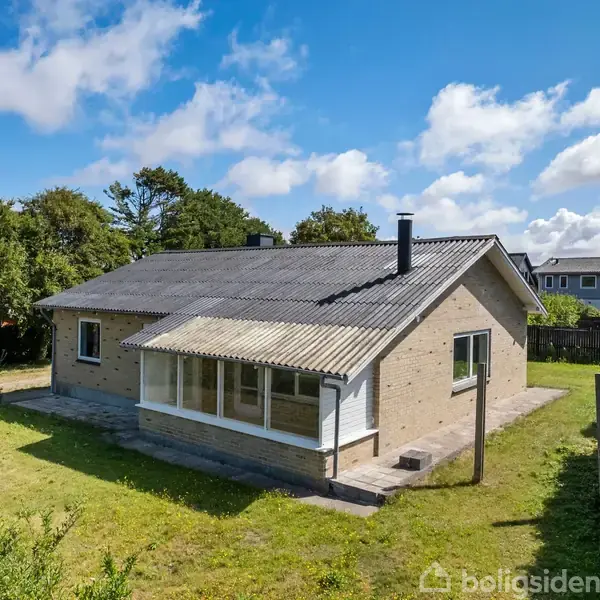 Et enfamiliehus med skråt tag og lille terrasse står omgivet af grønt græs og buske under en delvist skyet himmel.