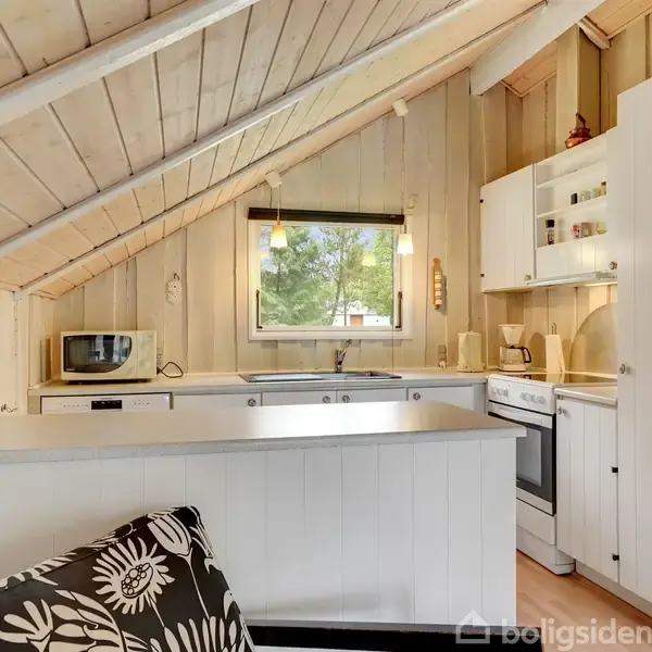 Et kompakt hvidt køkken med indbygget ovn, vask og mikroovn placeret under et skråt loft. En stor grøn vase med blomster står på bordpladen. Vindue med udsigt til træer.