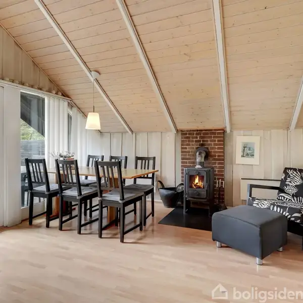 En stue med skrå loft, indeholder et spisebord med stole, en brændeovn i hjørnet, og en sofa med en skammel, belyst af naturligt lys fra store vinduer.