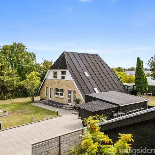 Et A-formet hus med mørkt tag og beige murstensvægge står i en grøn have med klare blå himmel i baggrunden. Plæne og træer omgiver ejendommen.