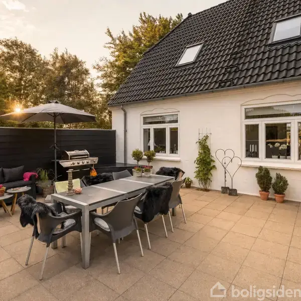 Et udendørs terrasseområde med et stort bord og stole dækket med tæpper, gasgrill ved siden af en sort væg og planter i potter. Hus i baggrunden med hvide vægge.