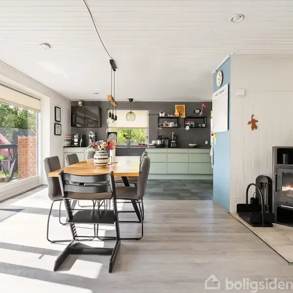 Spisebord med stole står i et moderne køkken-alrum. En kamin brænder til højre for bordet. Store vinduer giver udsigt til en have med murstensvæg og træer udenfor.